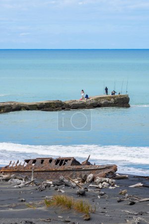 Foto de Patea Nueva Zelanda - 28 de febrero de 2024; Las personas que pescan en el extremo del rompeolas más allá del naufragio en la playa. - Imagen libre de derechos