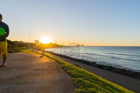 Foto de Nueva Plymouth Nueva Zelanda - 28 de febrero de 2024; vista frente al mar a lo largo de la costa hasta el sol poniente y la chimenea de la central eléctrica de Nueva Plymouth. - Imagen libre de derechos