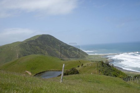 Hilly farmland with pond and view down to  Wairarapa coast and surf under blue sky