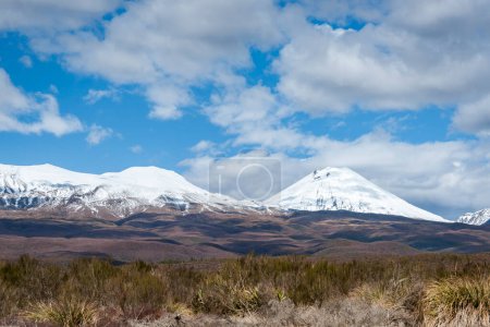 Clouds cast shadows over rolling landscape and snow-capped mountains in Tongariro National Park, New Zealand.