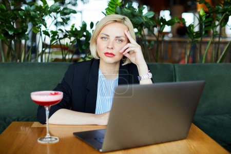 Bild eines inspirierten attraktiven Models, das an ihrem Laptop schreibt, Zeit allein im Café verbringt, ein Glas Cocktail auf dem Tisch hat, beschäftigt ist und Anzug trägt. Menschen und Tätigkeitskonzept.