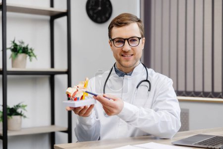 Foto de A young attractive otolaryngologist doctor is sitting at his workplace in the office and holding a model of the structure of the ear in his hands. The doctor consults the patient. - Imagen libre de derechos