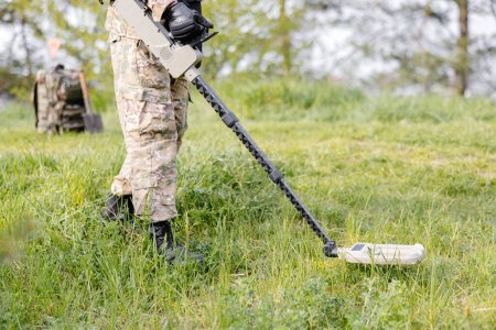 Un hombre con uniforme militar y chaleco antibalas trabaja en el bosque con un detector de metales. Un dragaminas realiza trabajos de desminado del territorio.