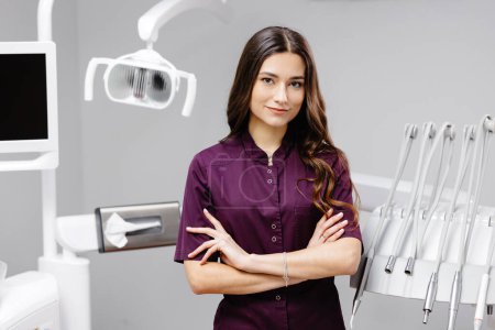 Photo for A young pretty female dentist is standing by the dental chair in the office with her arms crossed in front of her. - Royalty Free Image