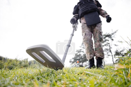 Photo for A man in a military uniform and bulletproof vest works in the forest with a metal detector. A minesweeper performs work on demining the territory. - Royalty Free Image