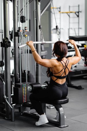 Photo for Muscular athletic female bodybuilder in black suit pulled on sports simulator in gym. Back muscles are very tense - Royalty Free Image