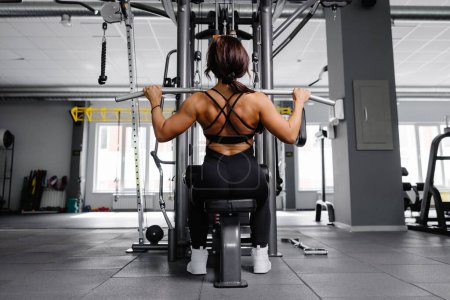 Photo for Muscular athletic female bodybuilder in black suit pulled on sports simulator in gym. Back muscles are very tense - Royalty Free Image