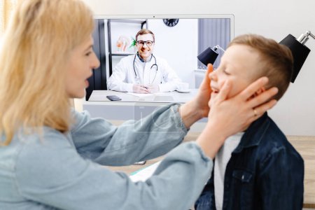 Photo for A young mother checks her son's throat while sitting at home and conducting an online doctor's consultation. Say aaah - Royalty Free Image