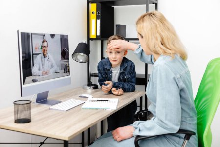 Mother checking son, touching hand on his forehead while attending video call with doctor over computer at home.