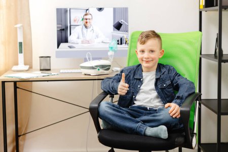 A cute smiling boy is sitting on a chair at home, and in the background is a table with a computer, at which a doctor is conducting an online consultation.