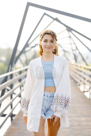 A woman wearing a white cardigan is standing on a bridge. She appears calm and relaxed as she gazes ahead.