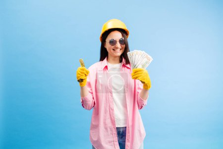 Young woman in yellow helmet holding cash money in banknotes isolated on blue background. Repair home concept.