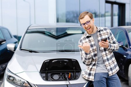 A young man uses a mobile phone while leaning on his electric car while the car is charging.