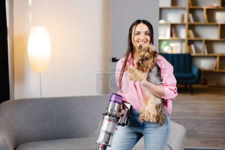Frau mit Staubsauger und Hund in der Hand staubsaugt im Wohnzimmer