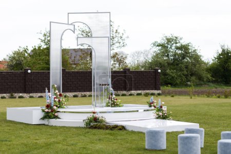 A white monument stands tall in a grassy area, adorned with colorful flowers. The contrast between the white structure and the vibrant flowers creates a striking visual impact.