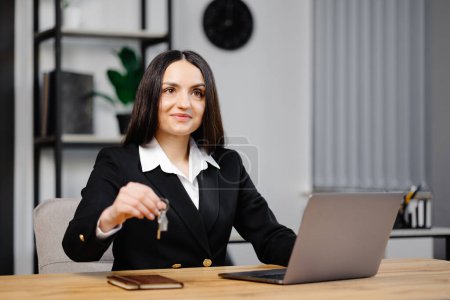 Young happy employee business woman wearing suit sitting at office desk with laptop and giving key in office.