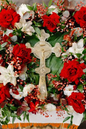 A cross stands prominently in the center of the scene, adorned with red and white flowers in full bloom. The contrast between the bright flowers and the solemn cross creates a striking visual impact.