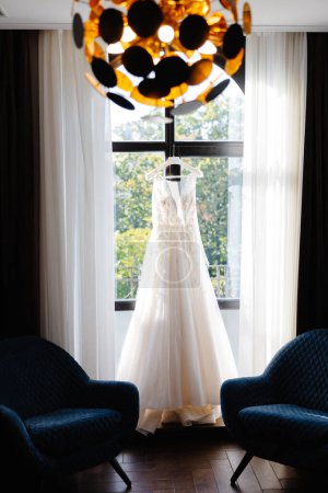 A white wedding dress hanging on a clothes rack in front of a window, with natural light shining through.