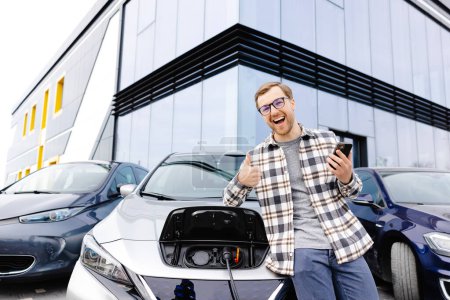 A young man uses a mobile phone while leaning on his electric car while the car is charging.