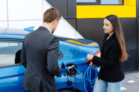 Gerente de ventas que muestra cómo cargar el coche eléctrico a una joven cliente, enchufando el cable en el enchufe del automóvil en el concesionario de automóviles al aire libre.