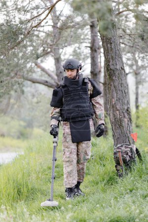 A man in a military uniform and bulletproof vest works in the forest with a metal detector. A minesweeper performs work on demining the territory.