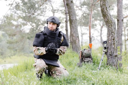 A man in a military uniform and a bulletproof vest works in the forest to demine the territory. A man puts on a protective helmet before starting work.