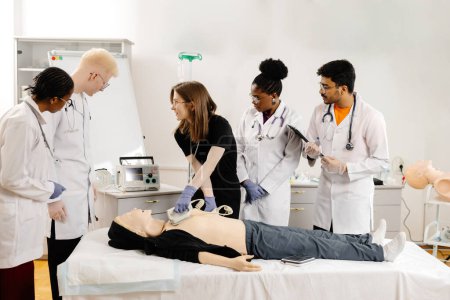 A group of medical students practice CPR on a mannequin in a classroom setting. A female student uses a defibrillator while others observe.