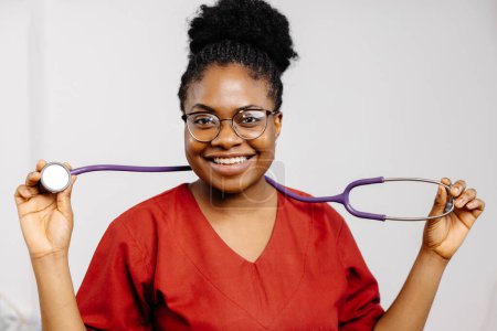 Eine lächelnde Ärztin im roten Peeling mit Brille hält ein Stethoskop in der Hand..