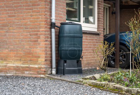 Foto de Barril de lluvia frente a una casa moderna, tanque de agua de lluvia para recoger el agua de lluvia y reutilizarlo en el jardín - Imagen libre de derechos