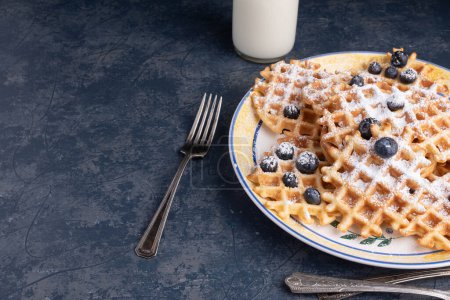 Photo for Waffles, blueberries, and powdered sugar on a blue tabletop with a glass of milk and linen napkin. - Royalty Free Image
