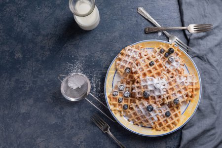 Photo for Waffles, blueberries, and powdered sugar on a blue tabletop with a glass of milk and linen napkin. - Royalty Free Image