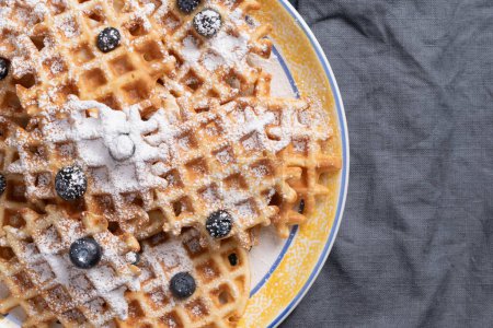 Photo for Waffles, blueberries, and powdered sugar on a yellow, blue, and white serving plate beside of a navy-blue linen napkin. - Royalty Free Image
