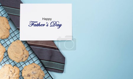 Feliz tarjeta del Día del Padre en corbata al lado de galletas recién horneadas de chispas de chocolate para papá. Fondo azul claro.