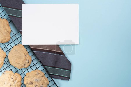 Corbata y tarjeta en blanco al lado de galletas fritas de chocolate recién horneadas. Fondo azul claro.
