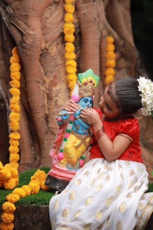 Une jolie petite fille enfant portant une robe Kerala couleur or jupe longue et chemisier rouge, assis sous un arbre banyan avec statue de seigneur krishna-fleur de douche dorée près de là