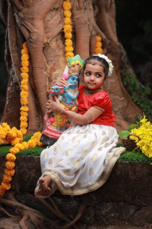 Téléchargez les photos : Une jolie petite fille enfant portant une robe Kerala couleur or jupe longue et chemisier rouge, assis sous un arbre banyan avec statue de seigneur krishna-fleur de douche dorée près de là - en image libre de droit