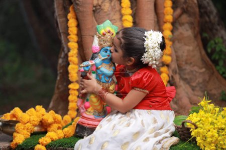 Una linda niña pequeña con Kerala vestido de color dorado falda larga y blusa roja, sentado bajo el árbol de Banyan con la estatua del señor krishna-flor de la ducha de oro cerca de