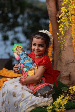 A cute small girl child wearing  Kerala dress-golden colour long skirt and red blouse, sitting under banyan tree with statue of lord krishna-golden shower flower near by