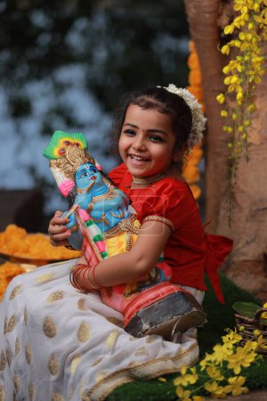 Photo for A cute small girl child wearing  Kerala dress-golden colour long skirt and red blouse, sitting under banyan tree with statue of lord krishna-golden shower flower near by - Royalty Free Image