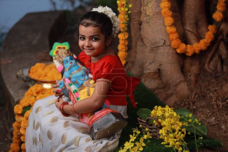Una linda niña pequeña con Kerala vestido de color dorado falda larga y blusa roja, sentado bajo el árbol de Banyan con la estatua del señor krishna-flor de la ducha de oro cerca de