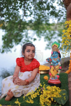 A cute small girl child wearing  Kerala dress-golden colour long skirt and red blouse, sitting under banyan tree with statue of lord krishna-golden shower flower near by