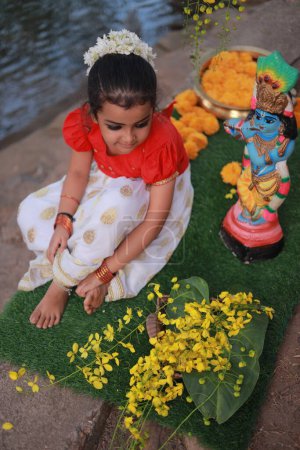 Téléchargez les photos : Une jolie petite fille enfant portant une robe Kerala couleur or jupe longue et chemisier rouge, assis sous un arbre banyan avec statue de seigneur krishna-fleur de douche dorée près de là - en image libre de droit