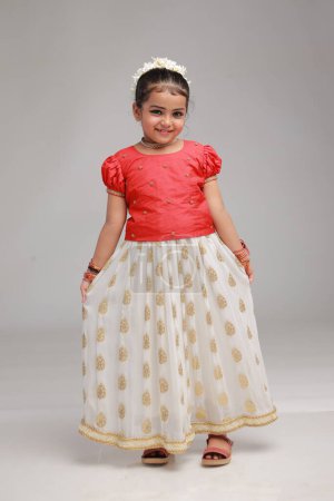 Une jolie petite fille enfant portant une jupe longue Kerala couleur or et chemisier rouge, image isolée avec fond blanc. 