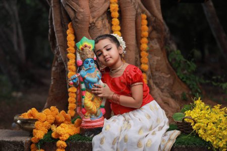 Foto de Una linda niña pequeña con Kerala vestido de color dorado falda larga y blusa roja, sentado bajo el árbol de Banyan con la estatua del señor krishna-flor de la ducha de oro cerca de - Imagen libre de derechos