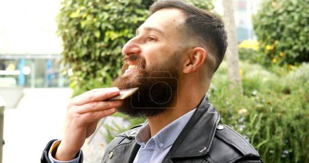 Téléchargez les photos : Jeune homme barbu se brossant la barbe en plein air. - en image libre de droit