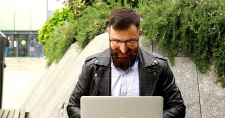 Foto de Joven barbudo trabajando en el ordenador portátil al aire libre. Hombre guapo escribiendo en el teclado portátil en la calle de la ciudad. - Imagen libre de derechos