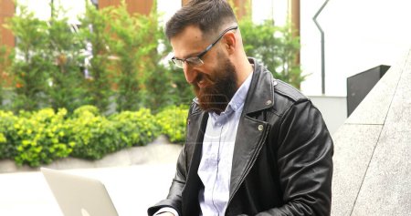 Foto de Joven barbudo trabajando en el ordenador portátil al aire libre. Hombre guapo escribiendo en el teclado portátil en la calle de la ciudad. - Imagen libre de derechos
