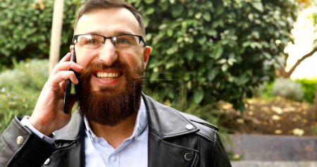 Téléchargez les photos : Beau jeune homme avec coiffure bien soignée et barbe à l'aide d'un smartphone extérieur. - en image libre de droit