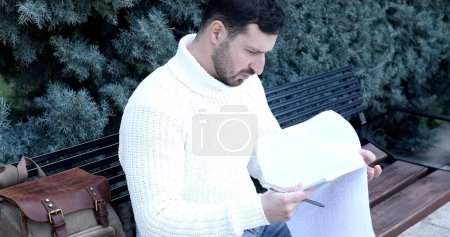Téléchargez les photos : Concentré jeune homme assis sur le banc et l'écriture dans le carnet dans le parc - en image libre de droit