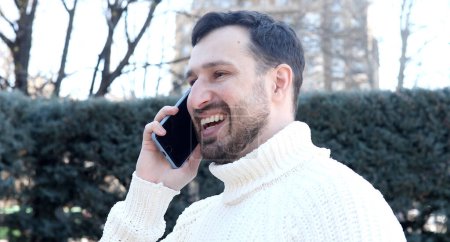 Téléchargez les photos : Émotionnel jeune homme parler par smartphone et rire en plein air - en image libre de droit